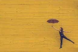 Umbrella in the sun; Lady jumping with umbrella in front of yellow wall